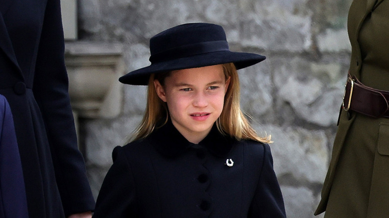Princess Charlotte wearing a black coat and hat and silver horseshoe brooch at Queen Elizabeth II's funeral