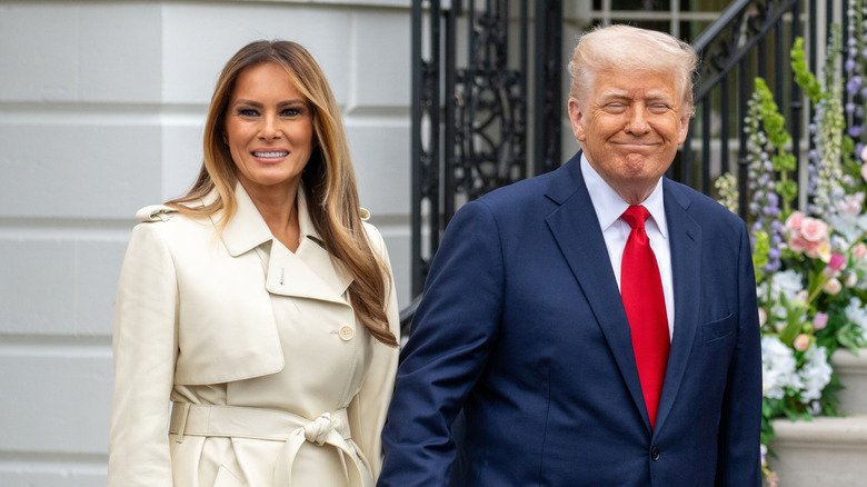 Melania Trump wearing a trench coat and Donald Trump wearing a suit outdoors as they both smile